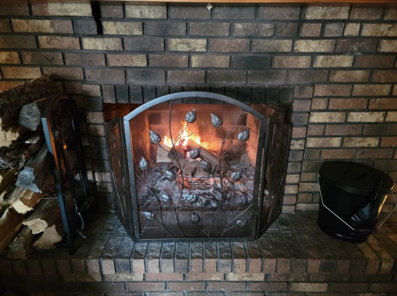 The fire place is made up of brown bricks with lots of chopped woods on its left and a black pale on its right. It has a design of metal leaves.
